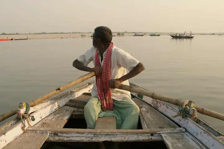 A boatman rows a wooden boat across the Ganga at Varanasi, with other boats on the river in morning light