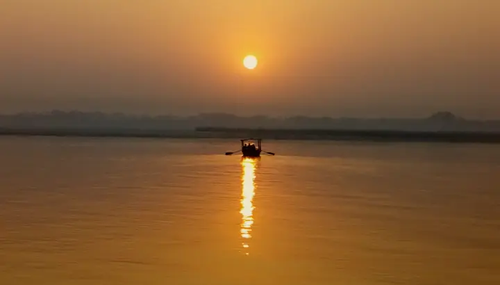 अस्सी घाट से गंगा पर सूर्योदय, सुनहरे पानी पर अकेली नाव की छाया।