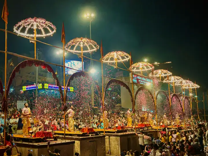 रात में दशाश्वमेध घाट पर गंगा आरती: जगमगाते छत्रों की कतार के नीचे मंचों पर पुजारी और पीछे श्रद्धालुओं की भीड़।